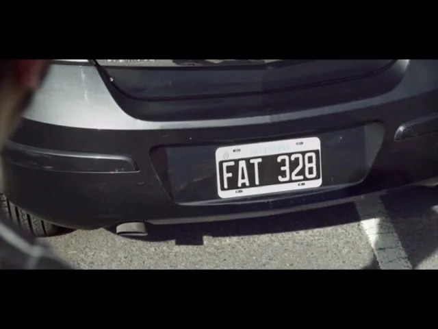 A person holds a cupcake while looking at a car's license plate that reads "FAT 328" in a parking lot.