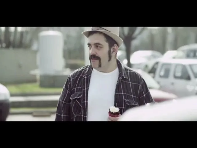 A man with a unique mustache and a fedora hat holds a cupcake, looking around a parking lot with cars in the background.