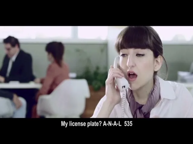 A young woman with bangs and a white shirt holds a phone to her ear, looking shocked while holding a document in her other hand, in an office.