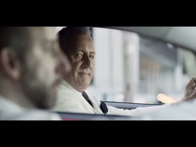 A man with a beard is driving a car, while a man in a white suit and black tie looks at him and smiles, in a city street.
