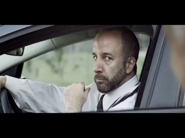 A man with a beard and white shirt sits in a car, looking over his shoulder at someone off-screen, with a serious expression.