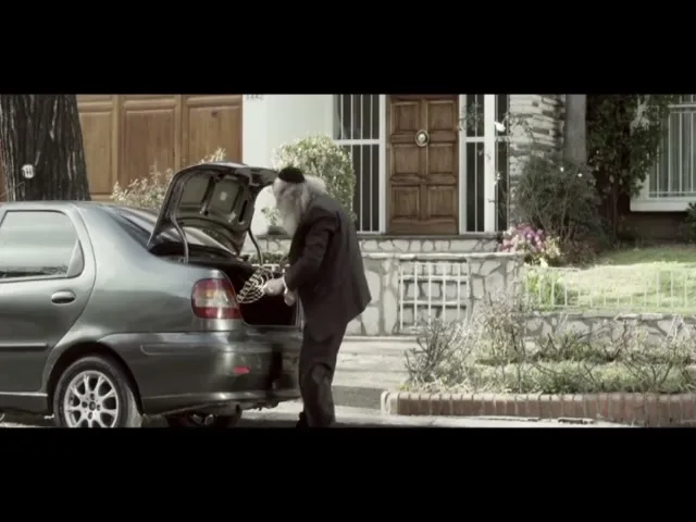 An elderly man with a long white beard and black suit struggles to close the trunk of a gray car in front of a house.