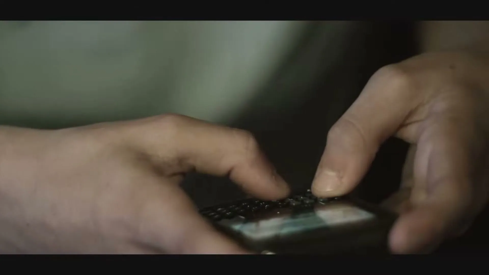 A person's hands are shown in a close-up, typing on a small, dark-colored mobile phone with a physical keyboard.