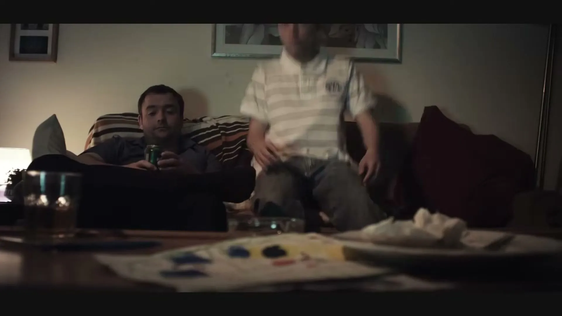 A man in his 30s drinks beer on a sofa while a teen boy sits next to him, both watching TV in a dimly lit living room.