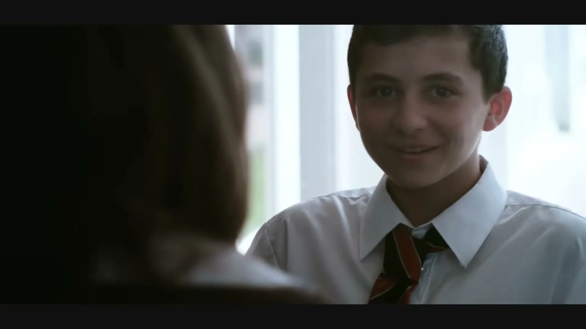 A young male student in a white shirt and tie looks at someone off-camera, with a window in the background.