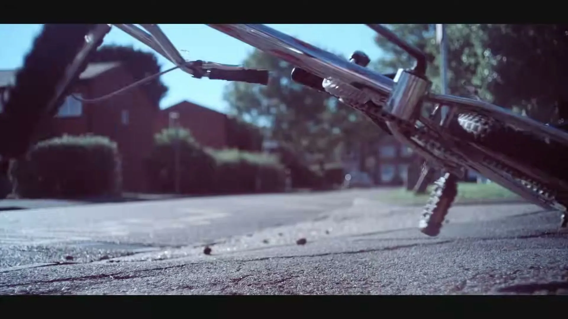 A bicycle falls to the ground on a suburban street on a sunny day, with houses and trees in the background.