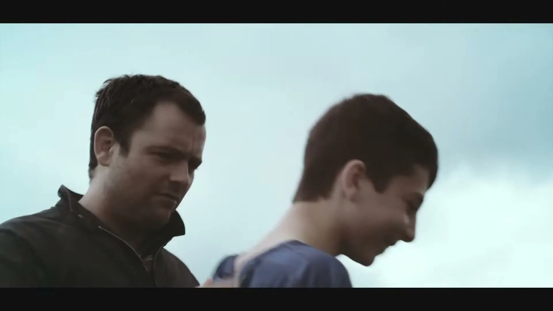 A man in a dark jacket puts his arm around a boy in a blue shirt, who then walks away, under a cloudy sky.