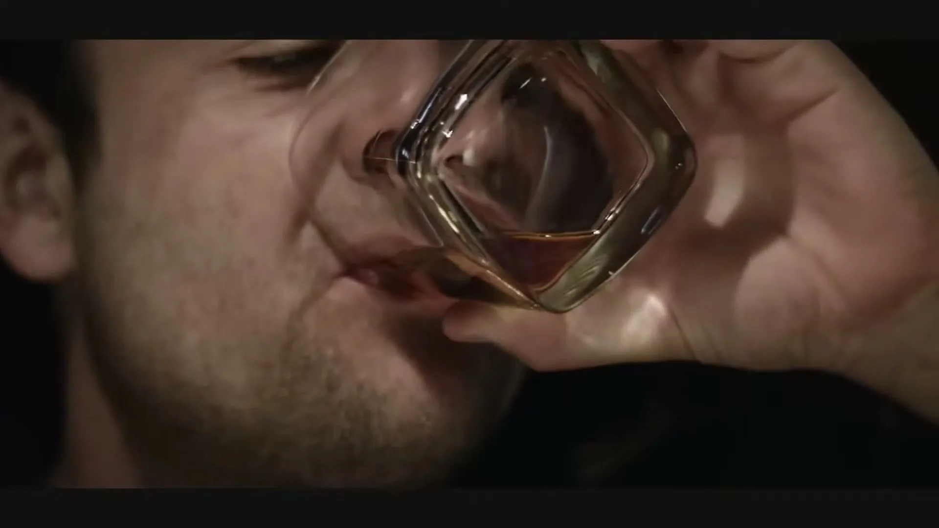 A man with stubble drinks from a glass of amber liquid, his face contorted in a grimace, in a dimly lit interior.