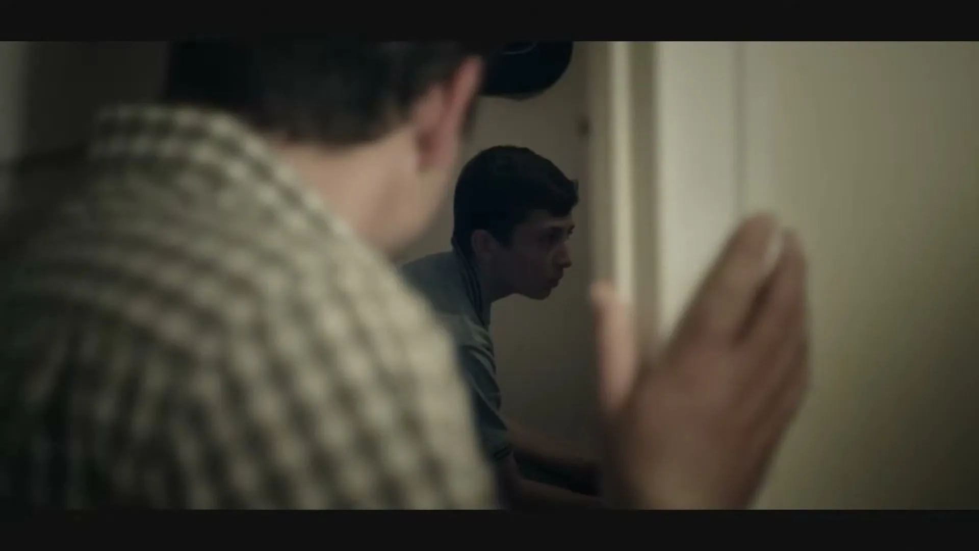 A young boy with dark hair, wearing a grey polo shirt, looks over his shoulder with a worried expression as an adult's back is seen in the foreground, in a dimly lit hallway.