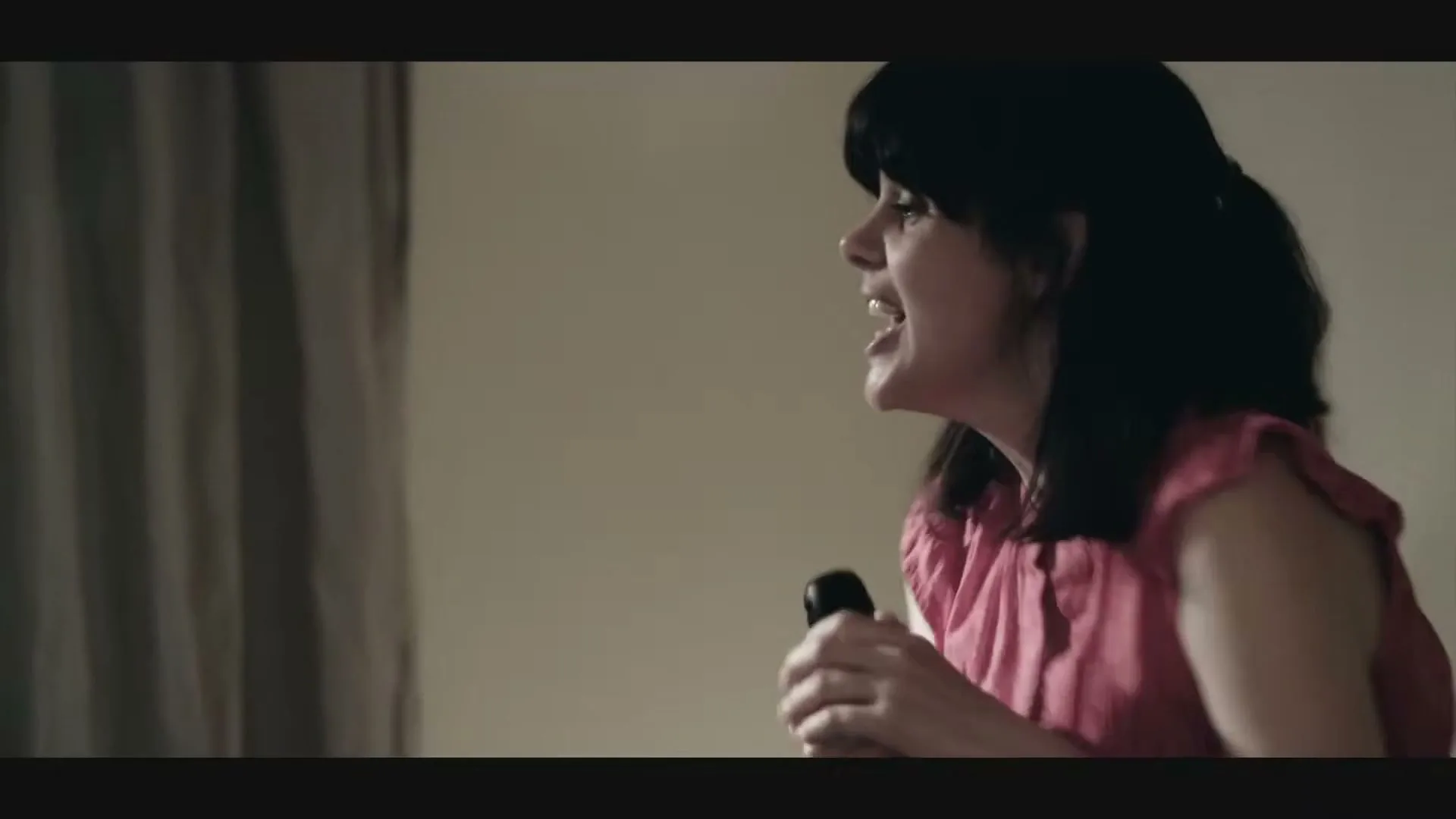 A young woman with dark hair, wearing a pink top, holds a microphone and sings passionately in an interior setting.