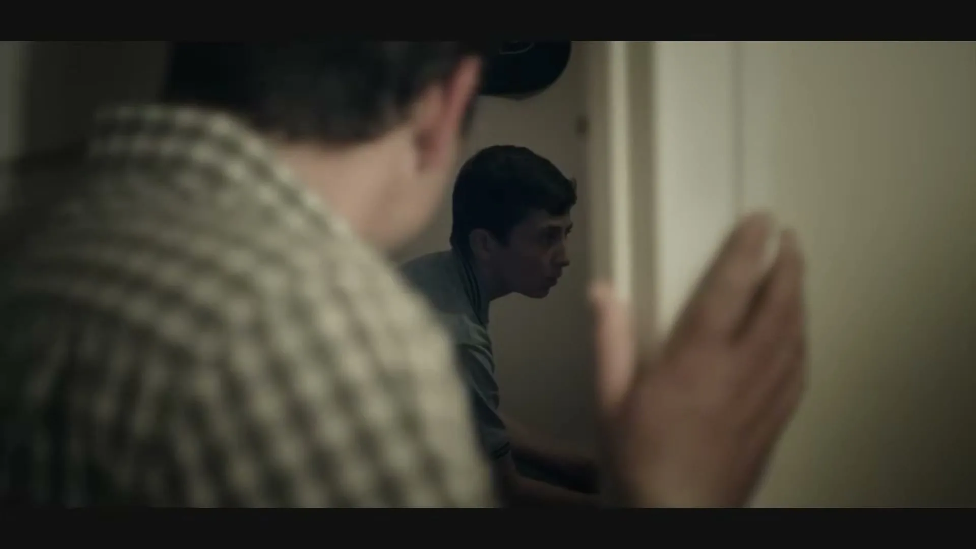 A young boy in a blue polo shirt looks fearfully from a doorway at an adult male's back in a dimly lit interior.