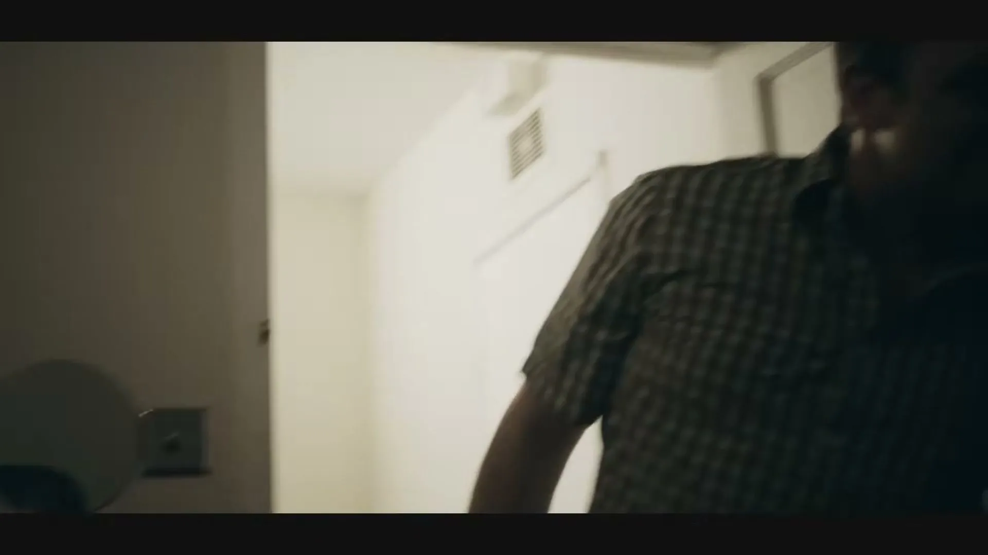 A man in a plaid shirt enters a room, looking at a younger person whose back is to the camera, in a dimly lit interior.