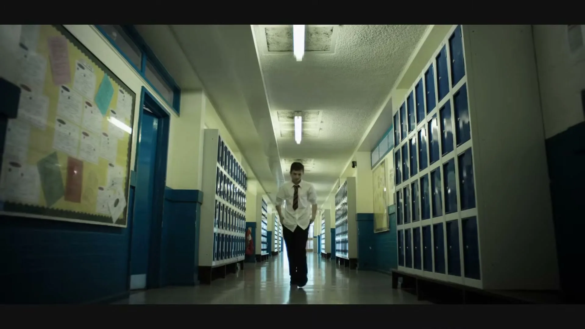 A male teen in a school uniform walks slowly down a long, empty school hallway lined with lockers, looking distressed.