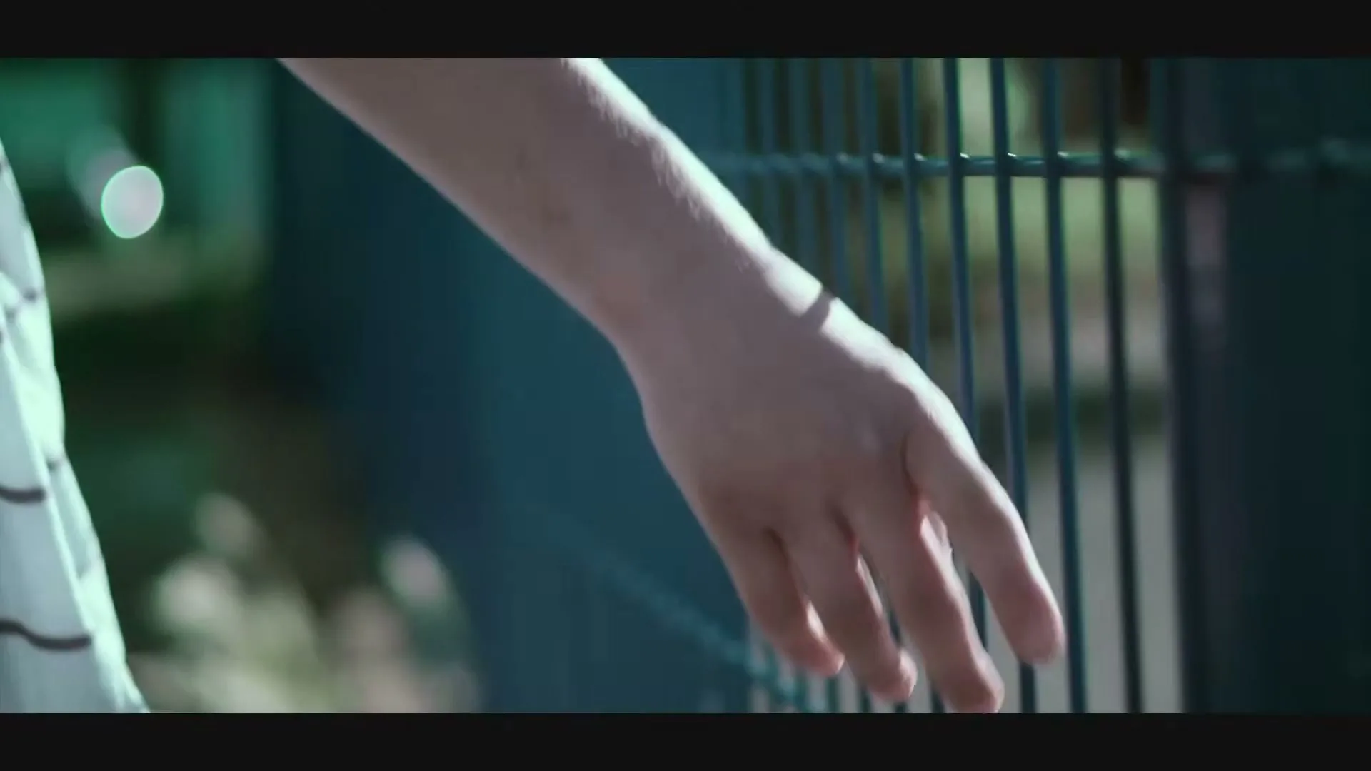 A child's hand with visible scars slides along a green metal fence in a park on a sunny day.