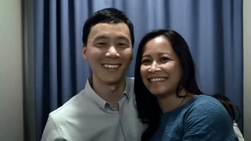 A smiling Asian couple poses for a photo in a photo booth, with the man's arm around the woman, against a blue curtain background.