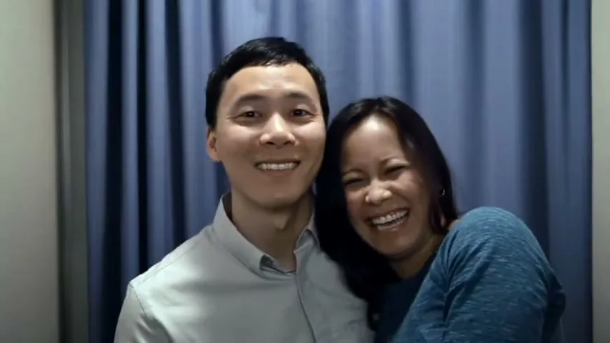 A smiling Asian couple, a man in a grey shirt and a woman in a blue top, pose for a photo in a photo booth with a blue curtain background.