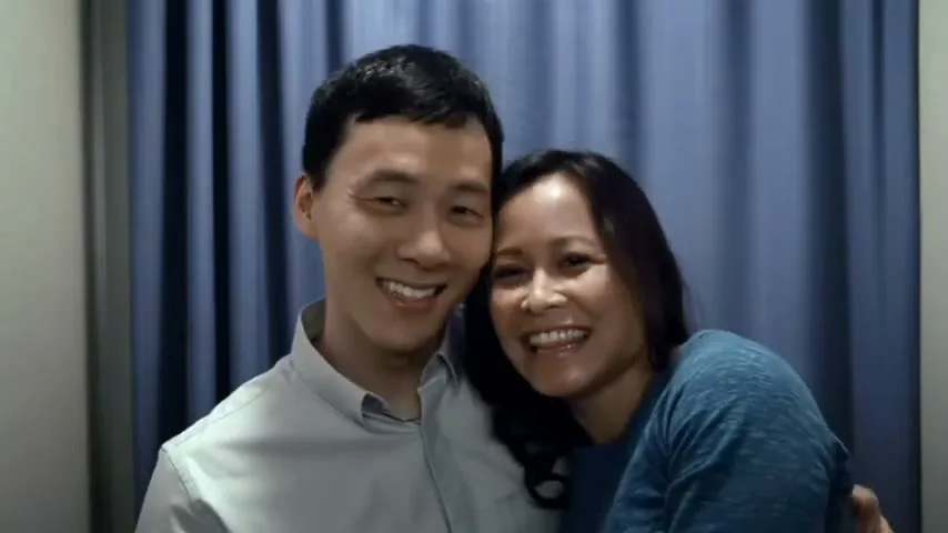 A smiling Asian man and a smiling Southeast Asian woman pose for a photo booth picture, with the woman leaning on the man's shoulder, in front of a blue curtain.