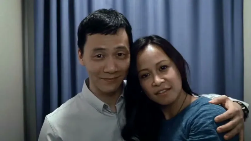 A smiling Asian man in a light shirt with his arm around an Asian woman in a blue shirt, who leans her head on his shoulder, in a photo booth.
