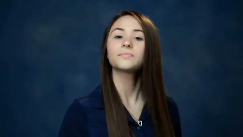 A young female with long brown hair and a blue shirt smiles at the camera, revealing braces, against a blue studio background.