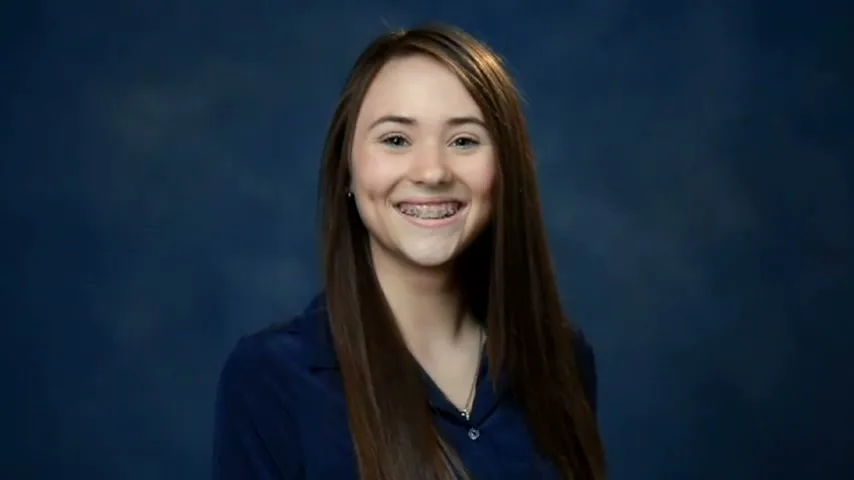 A young female with long brown hair and braces smiles at the camera in a school photo against a blue background.