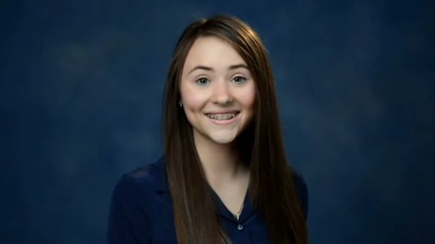 A young female with long brown hair and braces smiles at the camera in a school photo setting with a blue background.