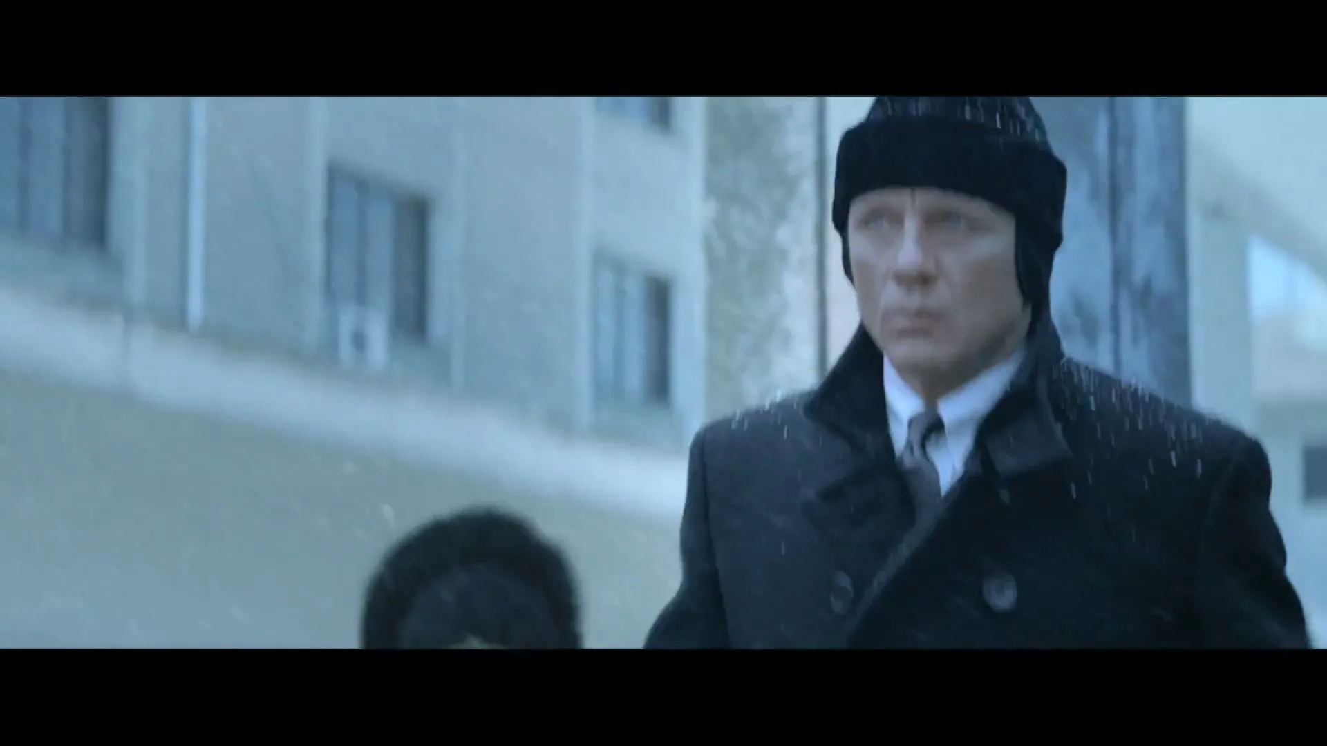 A man in a black coat and hat walks through a snowy city street, looking around cautiously as snow falls.
