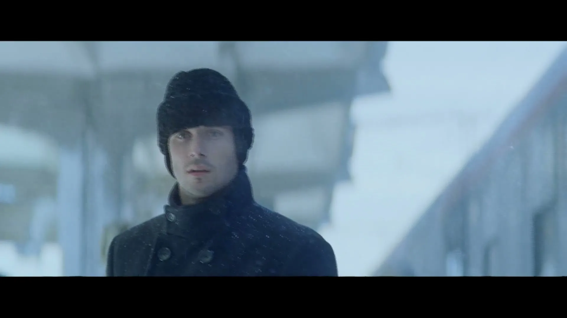 A man in a black coat and hat looks around nervously as snow falls on a train platform.