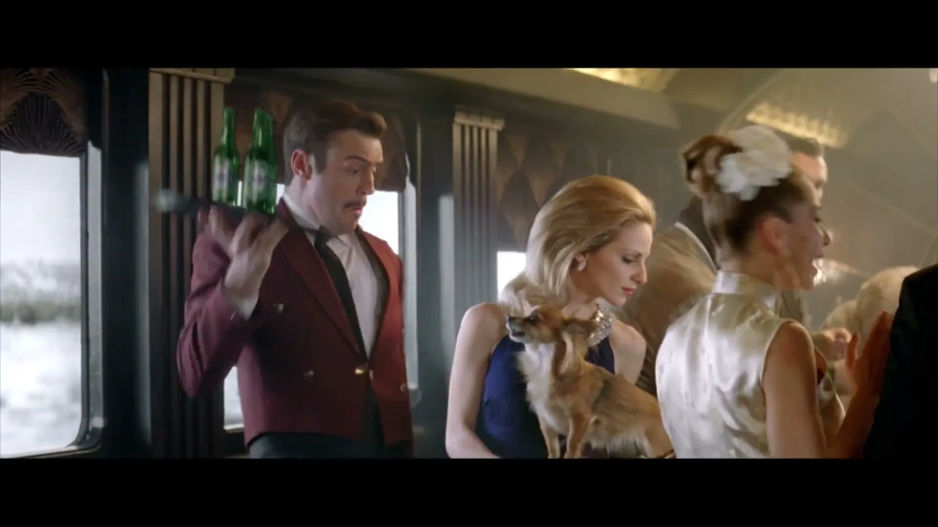 A waiter in a red jacket holds a tray of Heineken bottles while a woman in a blue dress holds a small dog on a moving train.