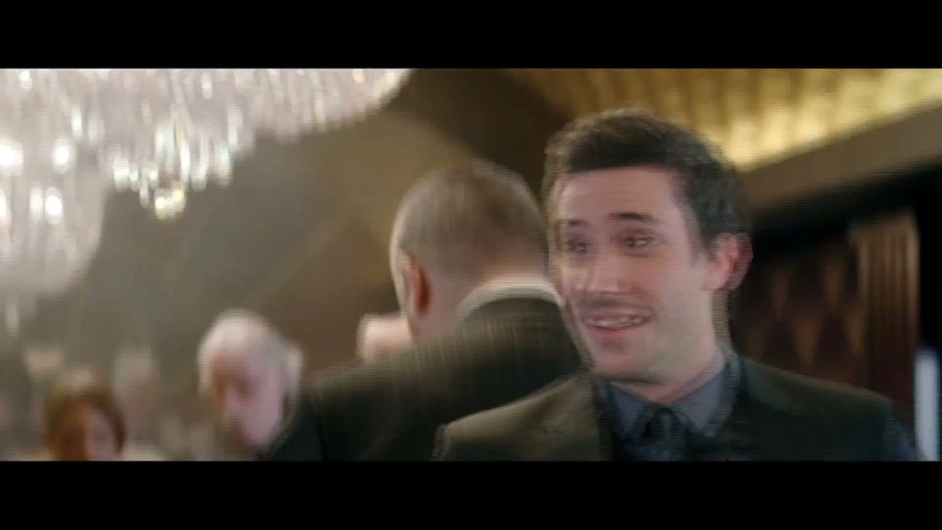 A young man in a suit looks surprised and then turns away in a crowded, elegant interior with chandeliers.