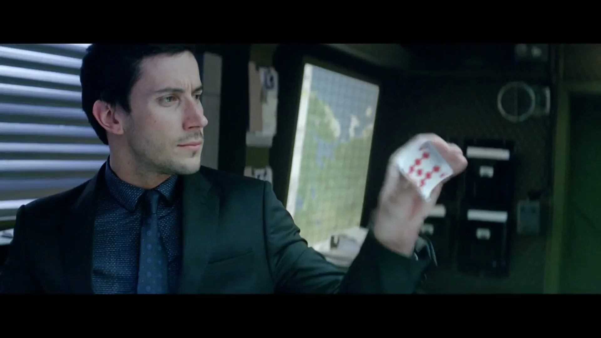 A man in a suit holds up a playing card, looking intently at it in a dimly lit room with a map on a screen.
