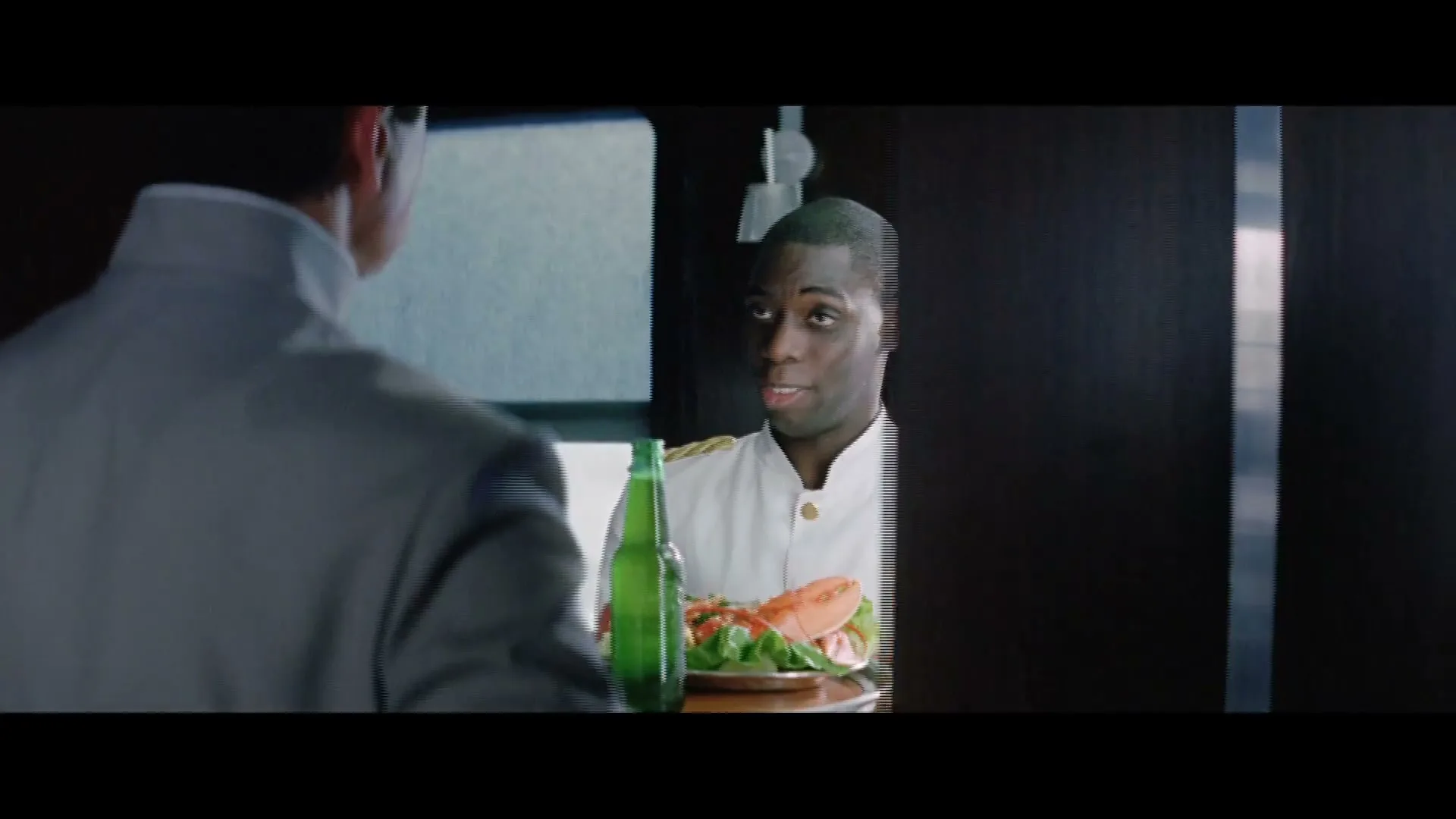 A man in a grey suit opens a door to reveal a waiter holding a tray with a lobster and a green bottle of Heineken beer.