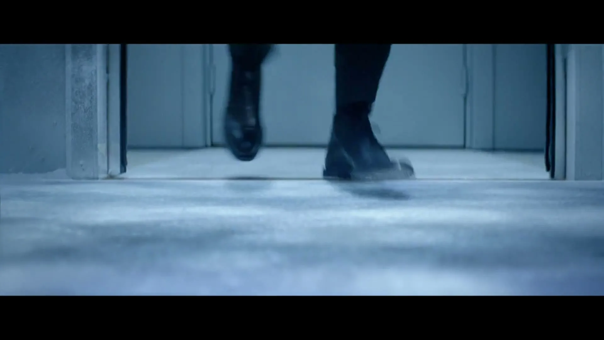 A man's feet in black shoes walk on a snowy, icy floor, kicking up snow as he moves forward in an interior space.