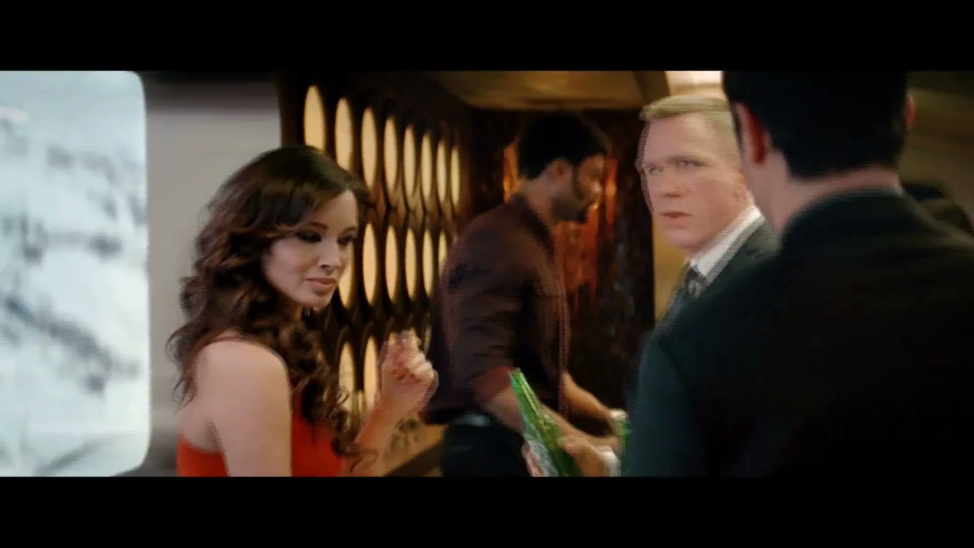 A man in a suit is handed a Heineken by a bartender, while a woman in a red dress watches, in a crowded bar.
