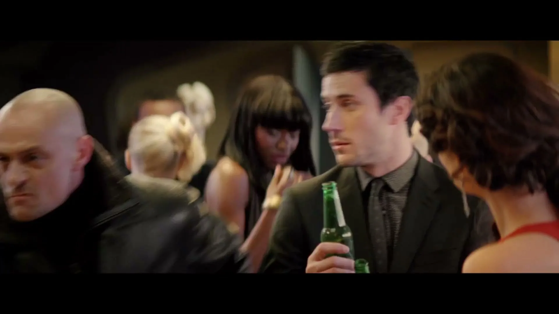 A man in a suit holds a green bottle, looking around a crowded, dimly lit room, then the camera pans down to a silver briefcase and a woman's legs in high heels.