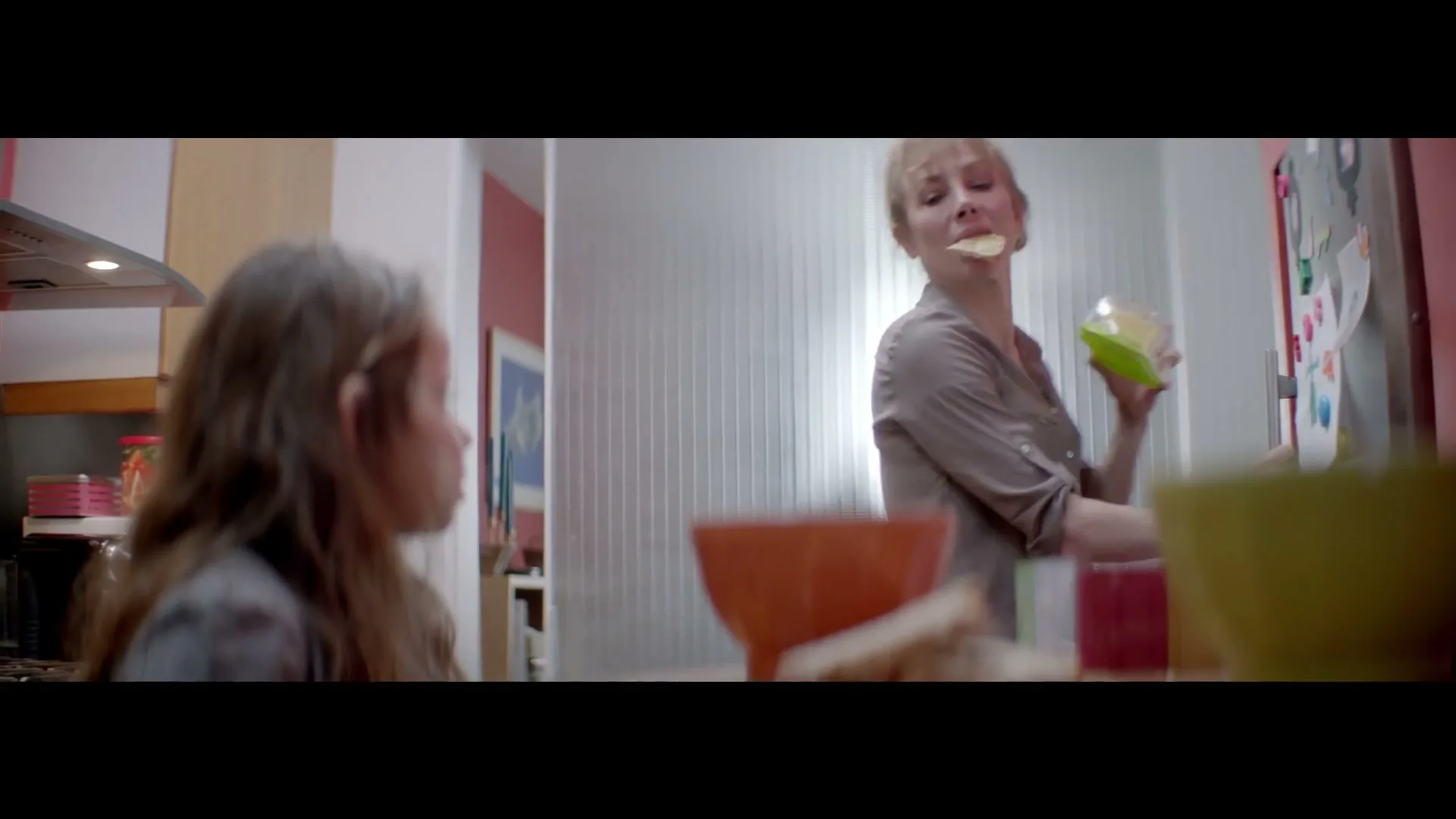 A young girl watches her mother, a woman in her 30s, playfully put a potato chip in her mouth while standing in a brightly lit kitchen.