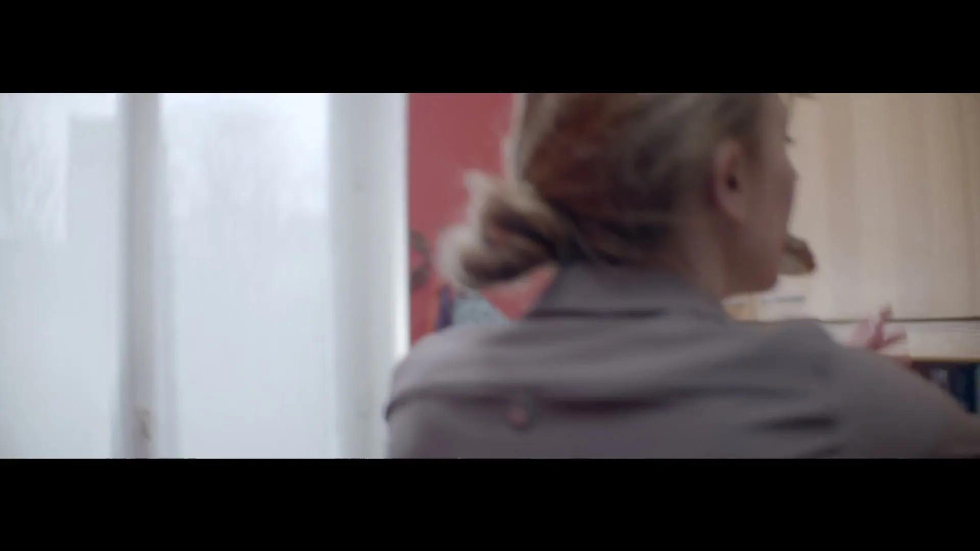 A woman with blonde hair in a bun, wearing a grey shirt, turns her head to the right in a kitchen.