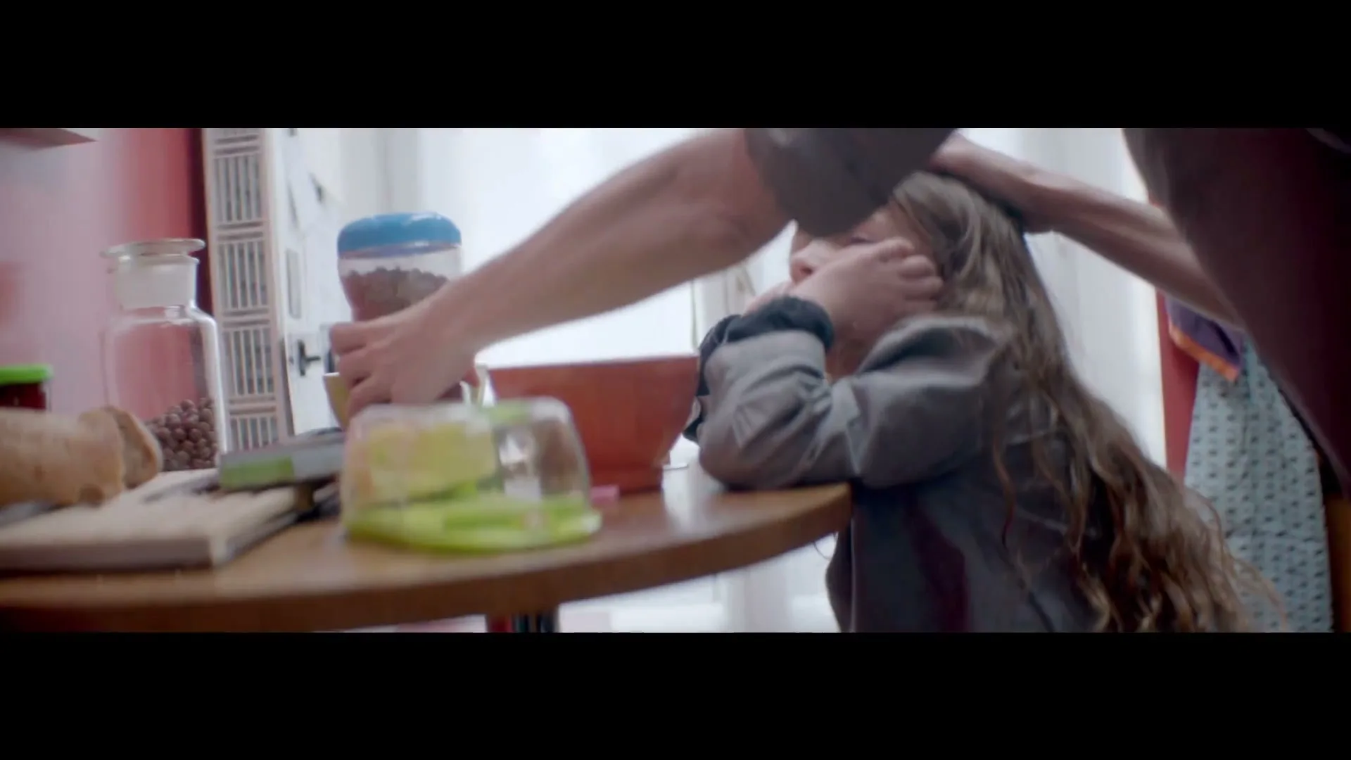 A young girl with long hair sits at a table, looking sad, while an adult's hands are on her head and another adult's hands pour cereal into a bowl.