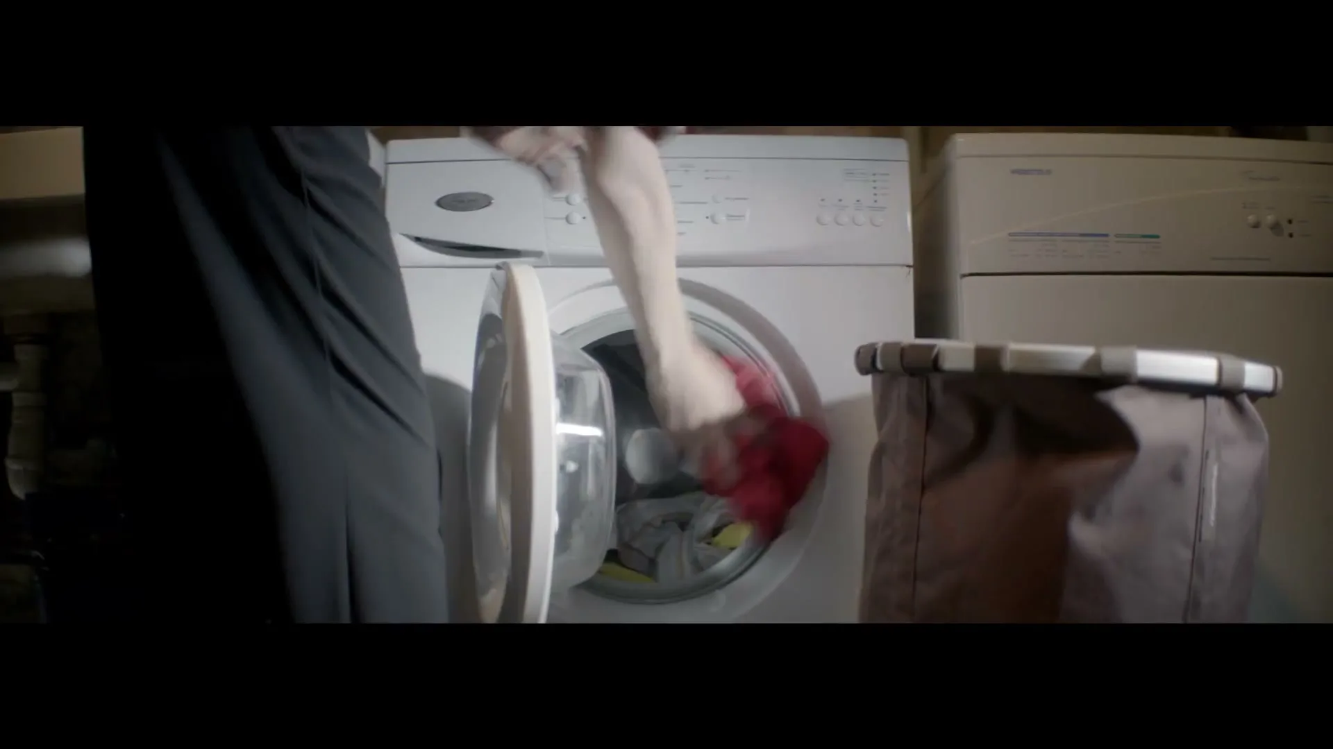 A person in a dark dress puts red laundry into a front-loading washing machine next to a laundry basket in a dimly lit room.