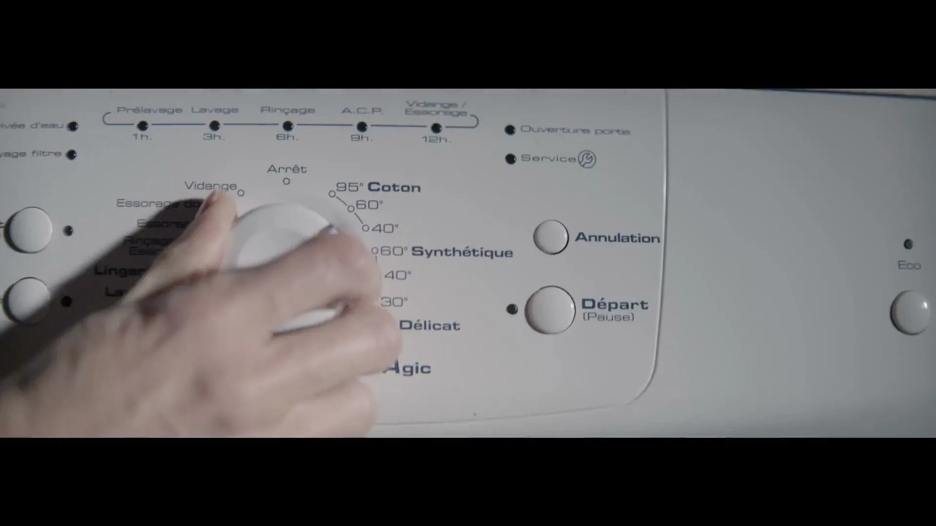 A person's hand turns the dial of a white washing machine, selecting a wash cycle in a dimly lit interior.