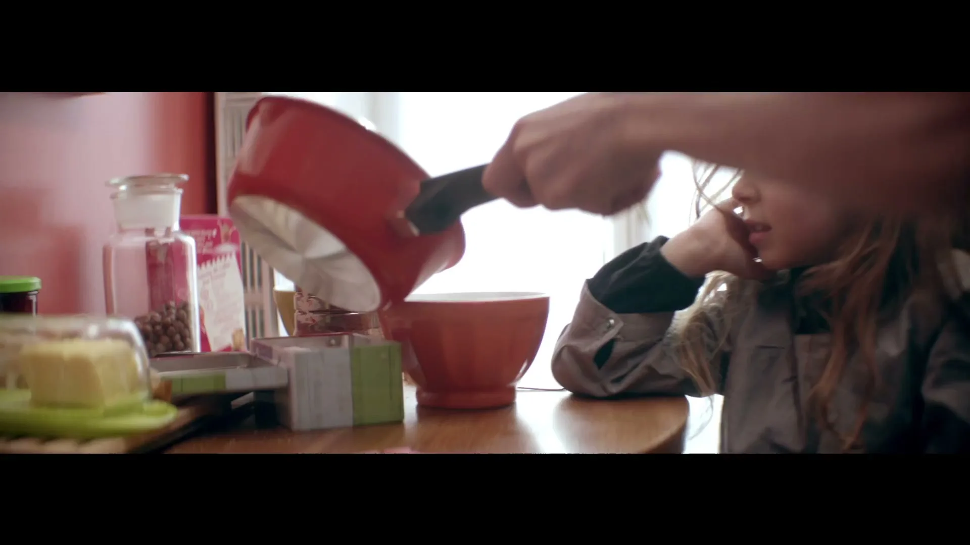 A person's hands pour liquid from a red saucepan into a bowl on a wooden table, next to a young girl with long hair.