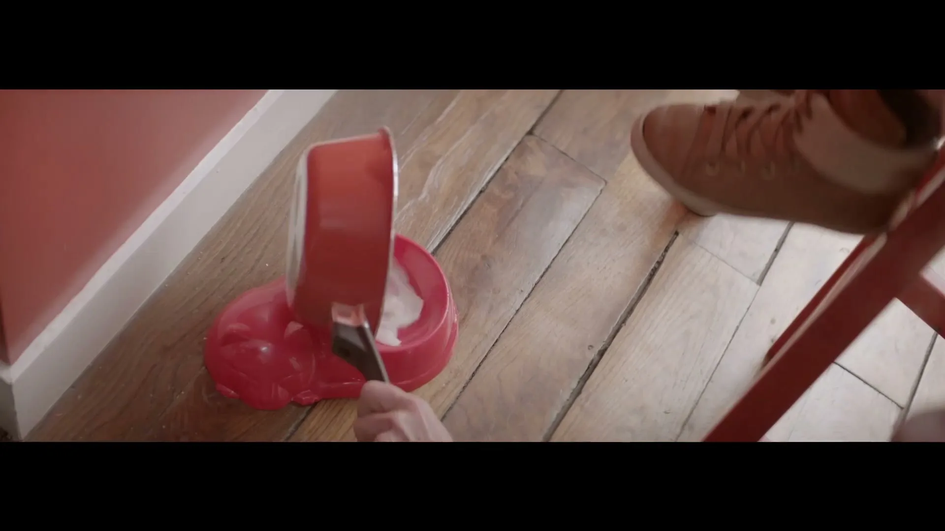 A person's hand pours food from a red pot into a red cat bowl on a wooden floor, with a person's foot visible.