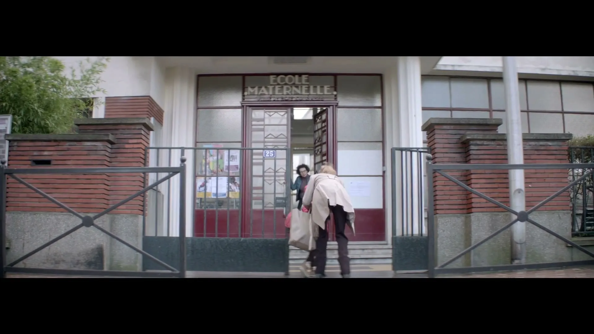 A woman in a trench coat and a child with a pink backpack walk out of a school building with "ECOLE MATERNELLE" written above the door.