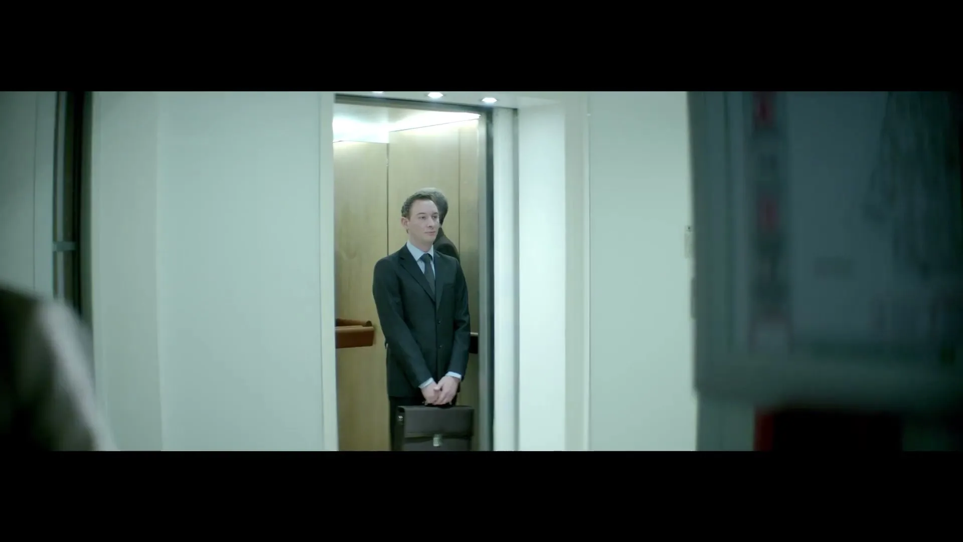 A woman in a trench coat walks past the camera, blurring the foreground, as a man in a suit stands in an elevator in an office building.