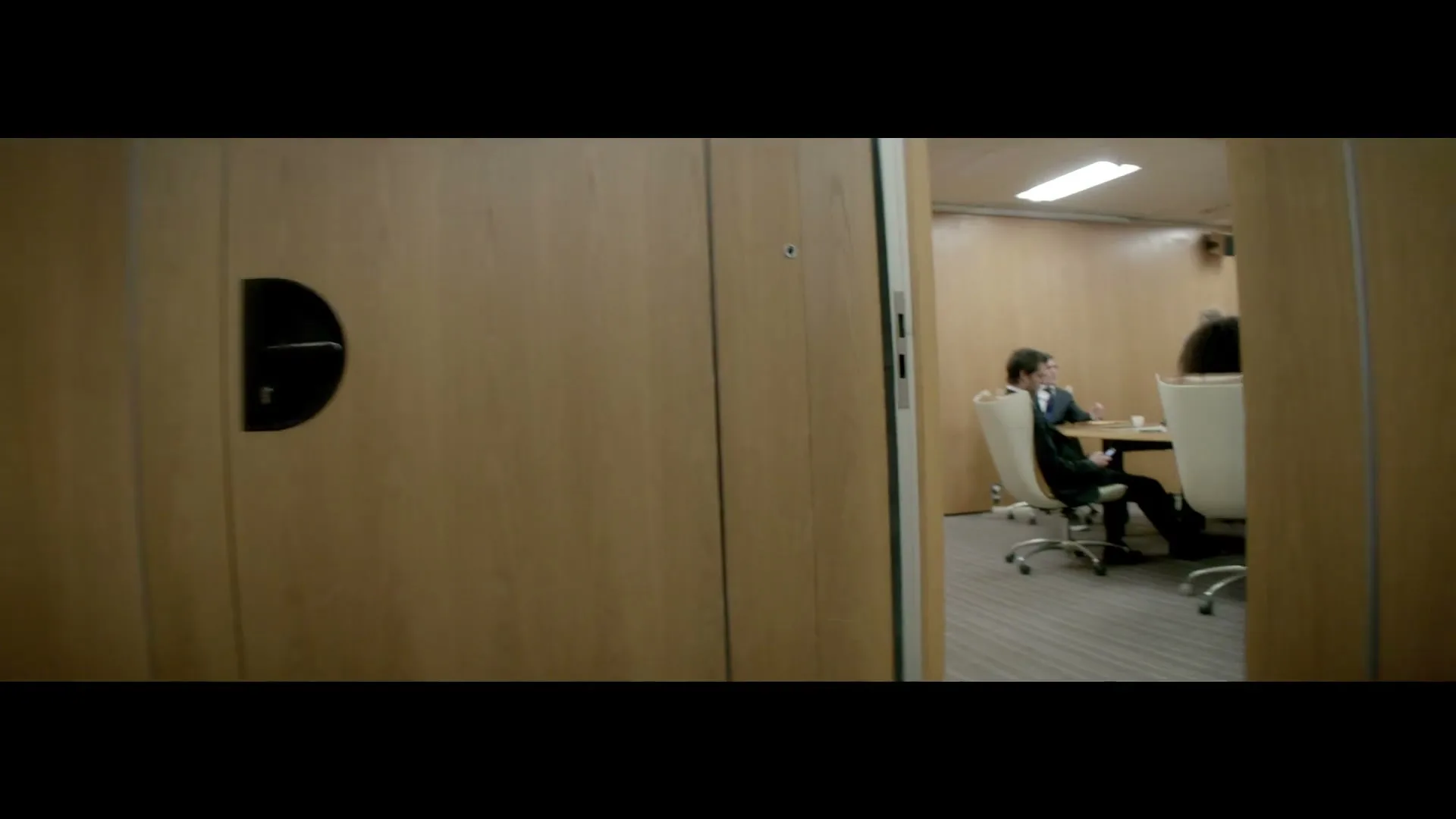 A camera moves past a wooden wall, revealing a glimpse into a modern office meeting room where several adults are seated around a table.
