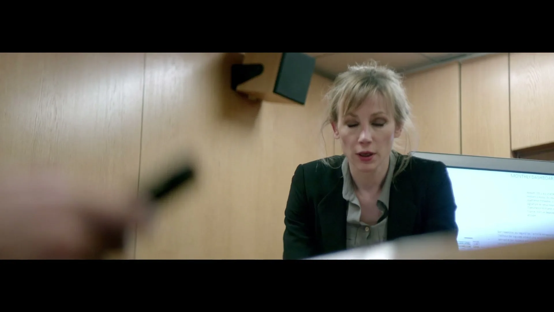 A blonde woman in a black blazer and grey shirt speaks to someone off-camera, with a blurred hand holding a pen in the foreground, in an office setting.