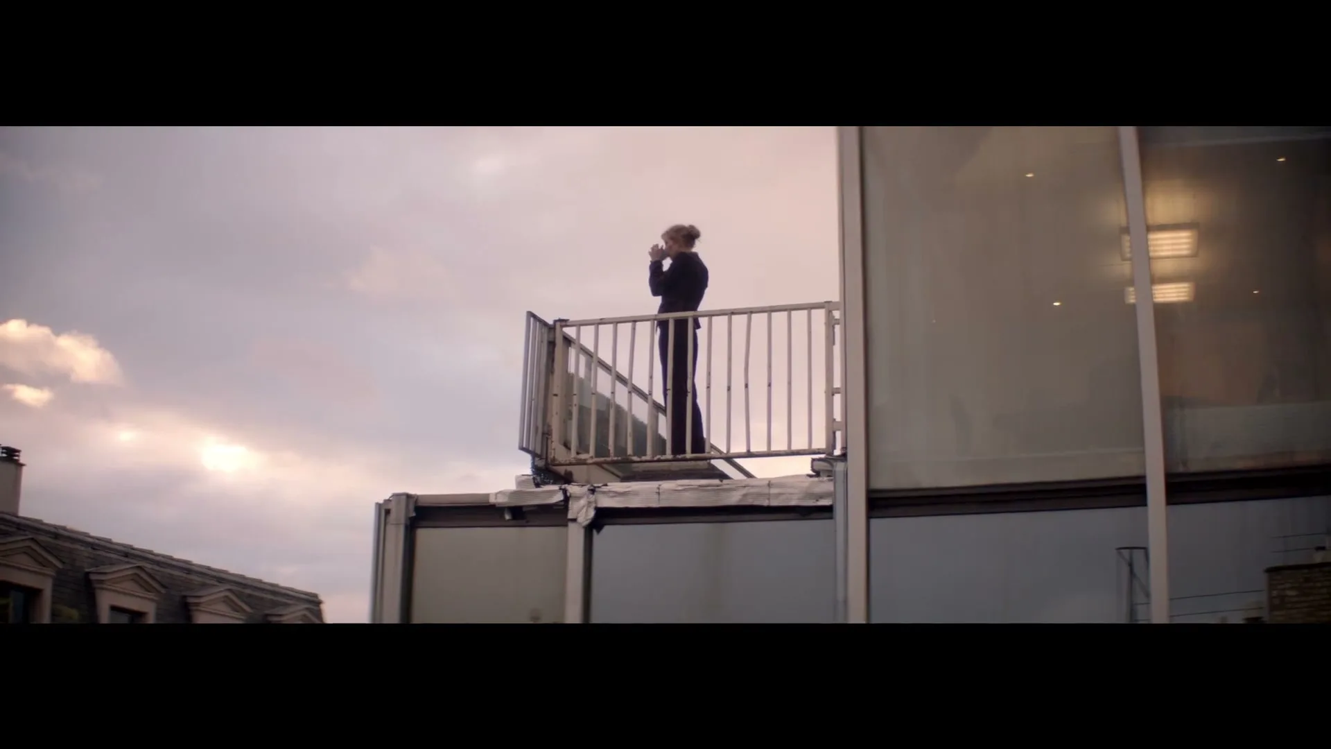 A woman in her 30s stands on a rooftop balcony, looking out at the city while holding a cup to her mouth, under a cloudy sky.