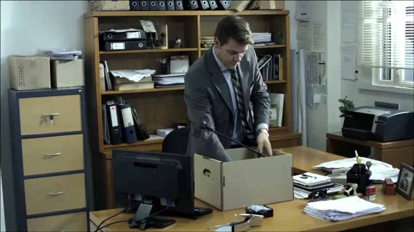 A man in a suit packs his belongings into a cardboard box on his desk in a messy office.
