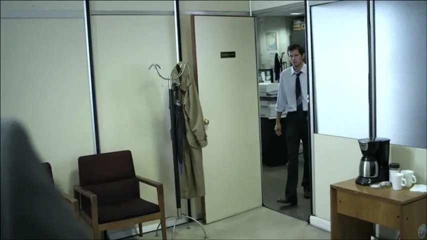 A man in a dress shirt and tie stands in an office doorway, looking into a waiting area with chairs and a coat rack.