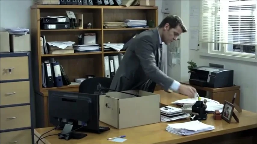 A man in a suit packs his belongings into a cardboard box on his office desk, surrounded by office supplies and a bookshelf.