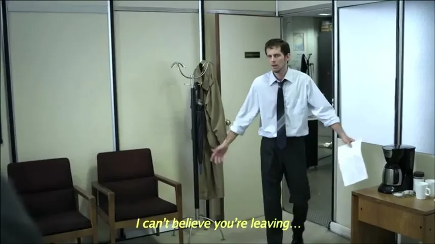 A man in a light blue shirt and dark tie walks into an office, holding papers, looking surprised at someone off-screen.